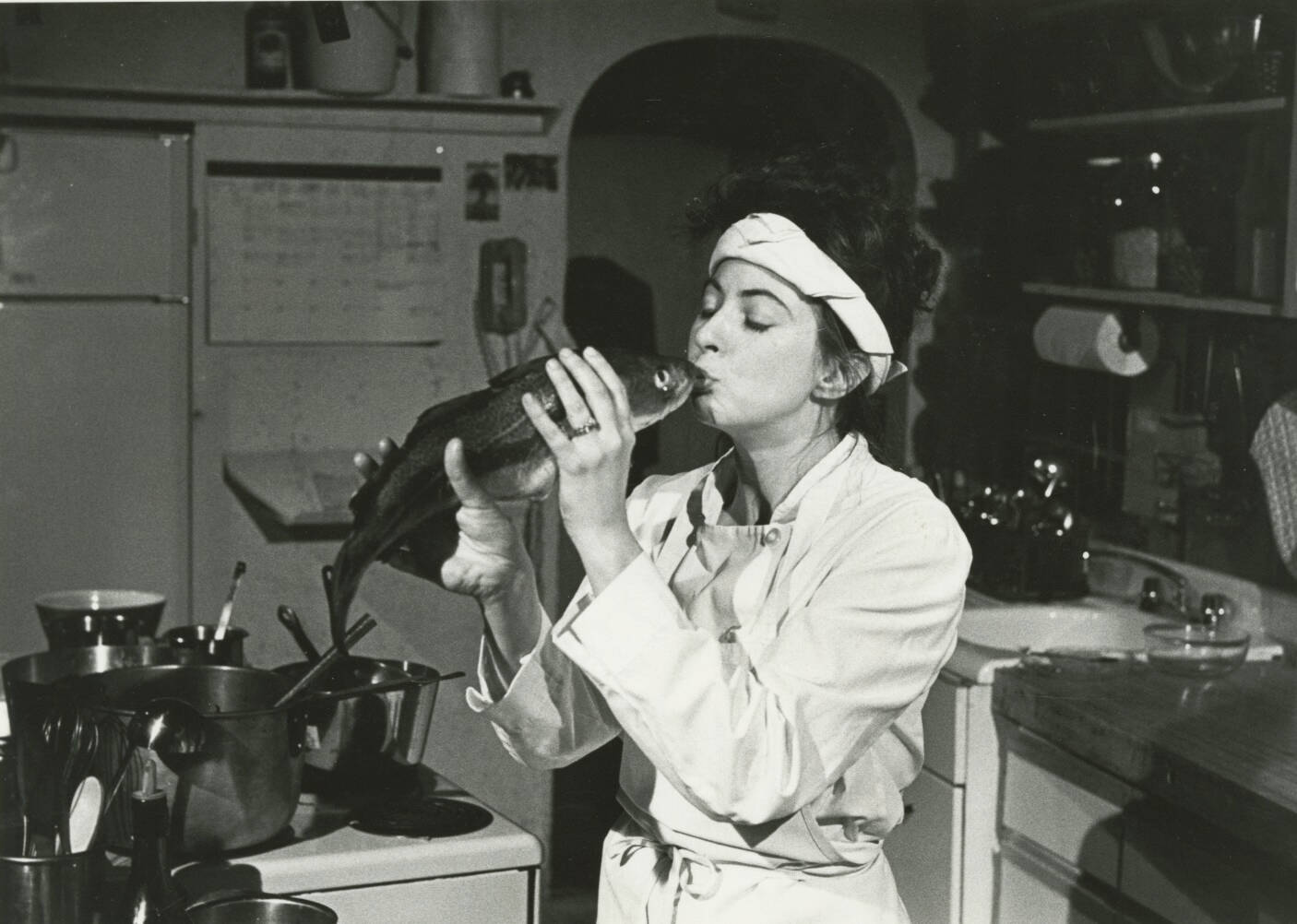 Black and white photograph of a woman in a kitchen kissing a rat.